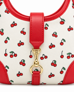 Close-up of Cherry Pattern Hamptons Hobo Bag showing gold horsebit snaffle clasp on center red faux leather strap tab, coated cream canvas with allover red cherry print and green stems, and red faux leather top trim with stitched edge detail
