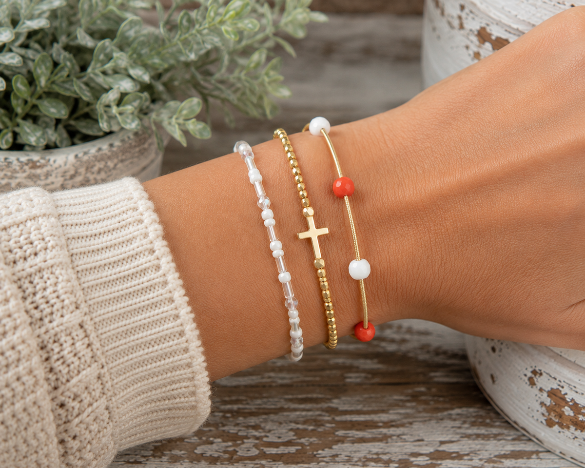 White iridescent seed bead bracelet stacked with gold cross beaded bracelet and red and white coil bracelet on wrist with cream knit sweater and rustic white pedestal backdrop
