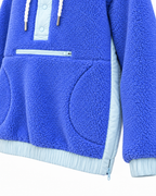 Close-up of the blue sherpa hoodie showing the front zippered pocket, curved hand-pocket opening, light blue side panel, and side-hem zipper
