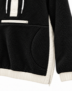 Close-up of the black sherpa hoodie showing the front zippered pocket, curved hand-pocket opening, ivory side panel, and contrasting cuff and hem
