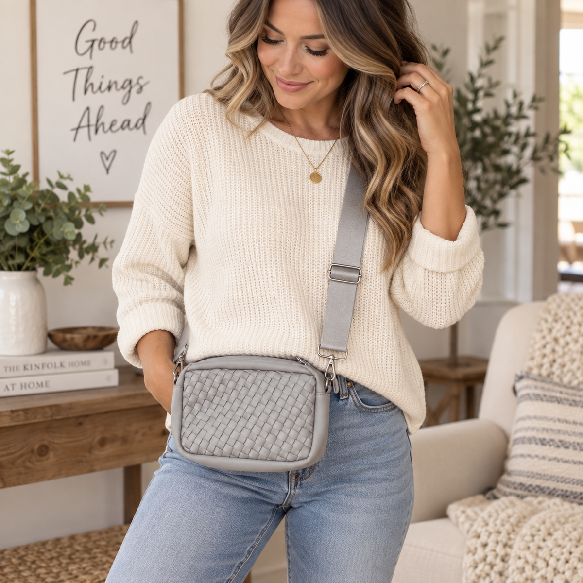 Woman wearing Gray Woven Crossbody Bag across body showing light gray interlaced woven panel, smooth gray trim, and adjustable wide gray webbing strap with cream sweater and jeans in bright farmhouse living room with Good Things Ahead sign
