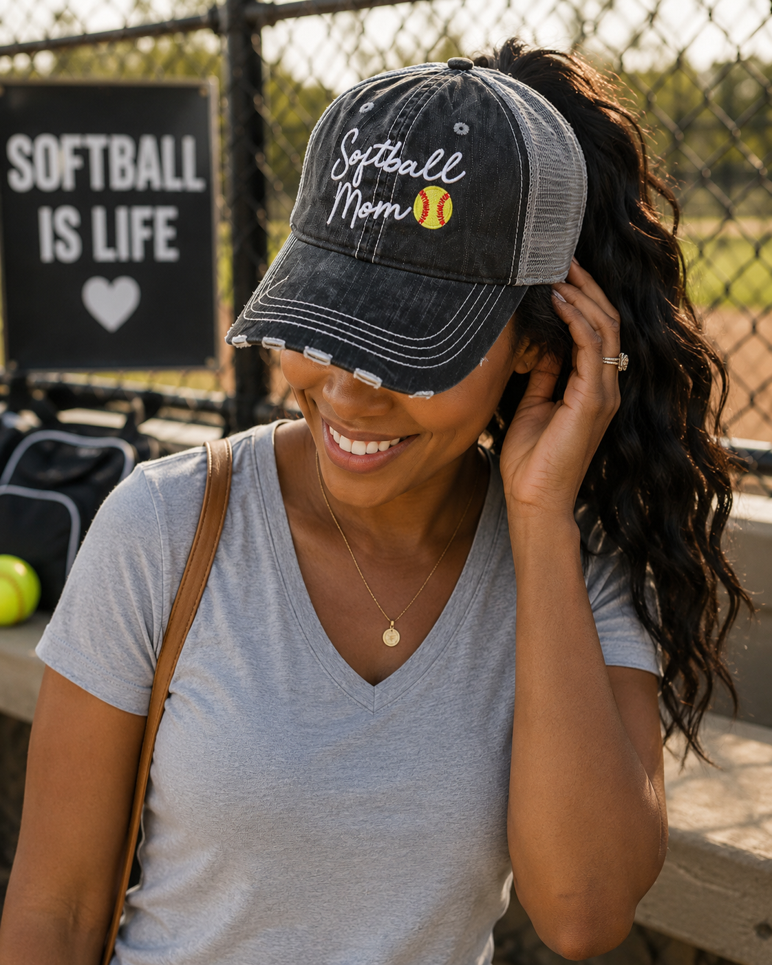 A woman wearing a dark gray distressed baseball cap with 'Softball Mom' and a softball graphic embroidered on the front, photographed outdoors at a softball field.