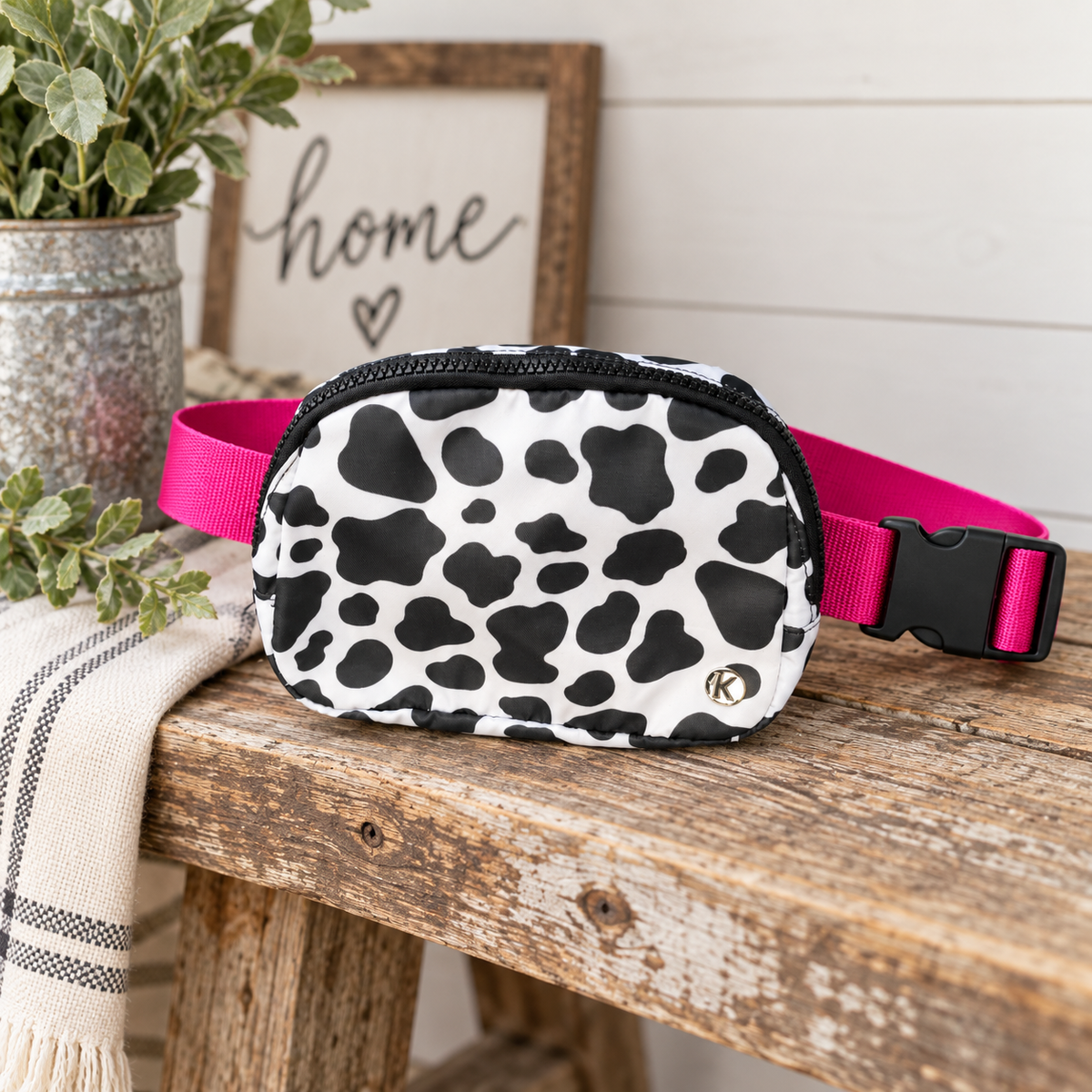 Cow Print Belt Bag on rustic wood bench showing white nylon with large black cow print spots, silver Katydid K logo rivet, and hot pink webbing strap with black side-release buckle beside galvanized metal vase of eucalyptus greenery, cream plaid linen throw, and framed Home sign on white shiplap background
