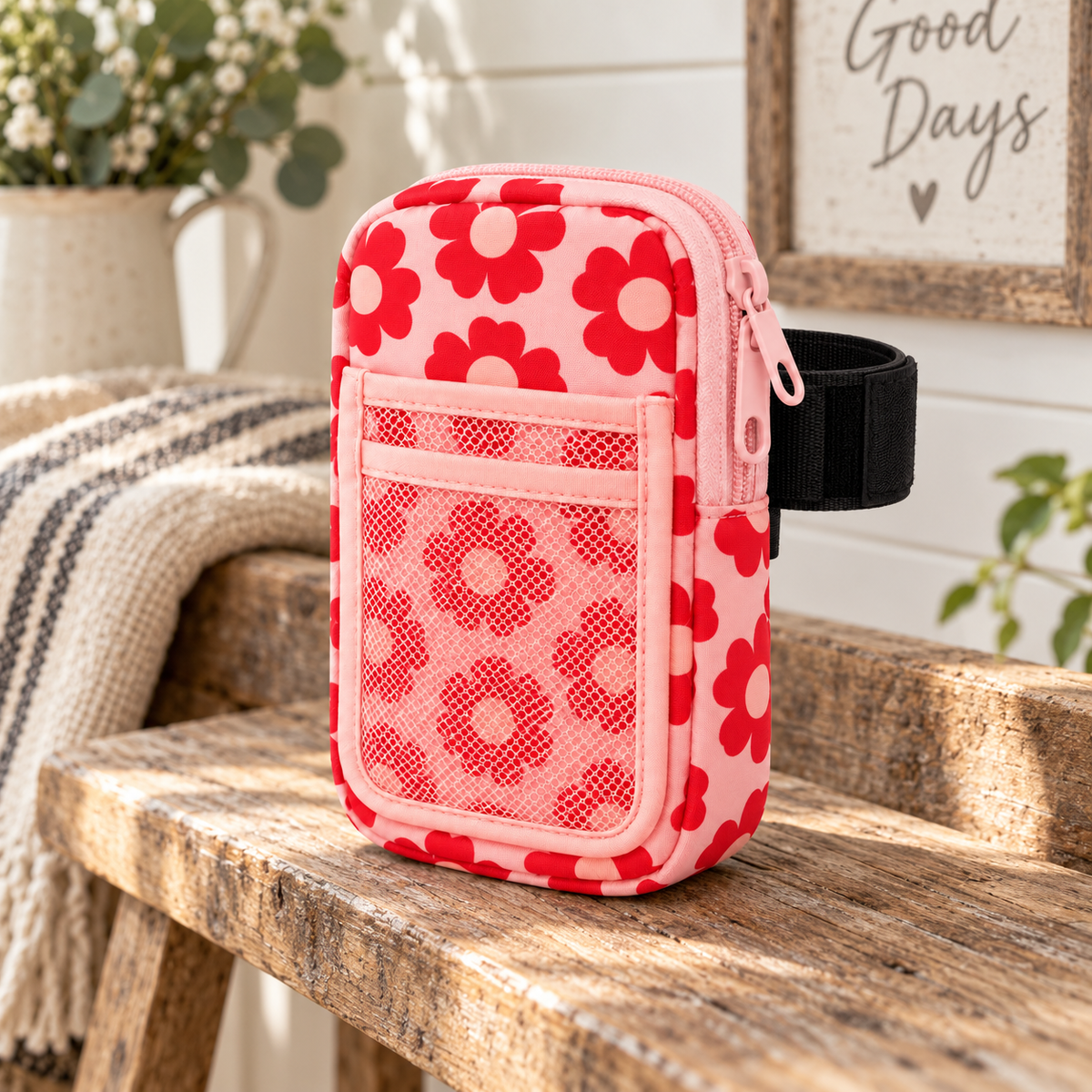 Pink Retro Floral Tumbler Pouch on rustic wooden bench with black elastic tumbler strap showing bold red retro daisy print, pink top zipper, slip pocket, and mesh pocket beside striped throw blanket, white pitcher, and Good Days sign with greenery
