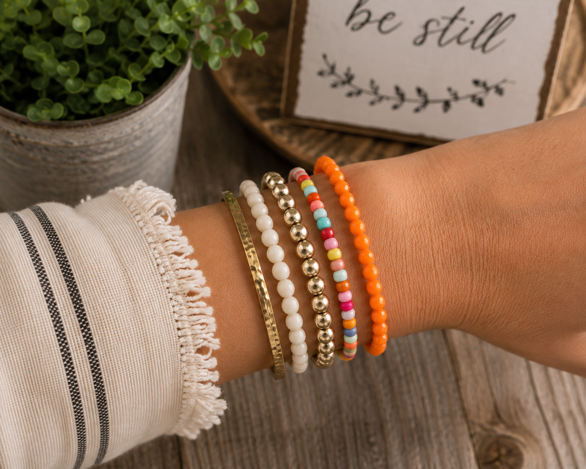 Orange faceted bead bracelet stacked with colorful seed bead bracelet, gold ball bracelet, white bead bracelet, and hammered gold bangle on wrist
