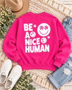 Back of the hot pink Be a Nice Human sweatshirt styled with jeans, white sneakers, a tan hat, flowers, and greenery
