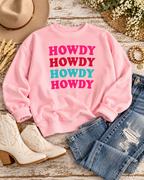 Light pink Howdy sweatshirt styled with distressed jeans, cream Western boots, a tan hat, bracelets, and flowers on rustic wood
