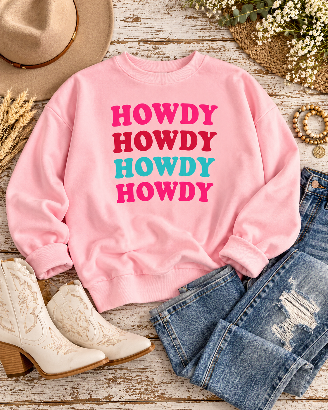 Light pink Howdy sweatshirt styled with distressed jeans, cream Western boots, a tan hat, bracelets, and flowers on rustic wood
