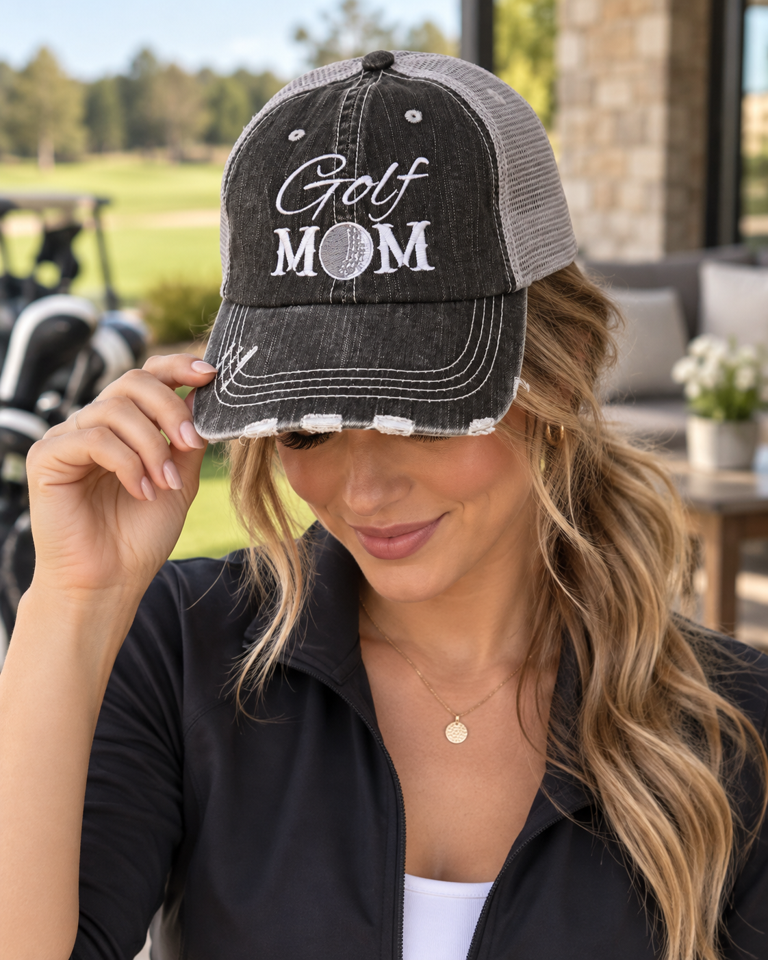 Woman wearing Golf Mom Trucker Hat with distressed black front and gray mesh back showing white embroidered Golf Mom script and block lettering with golf ball detail with black zip jacket and golf course backdrop
