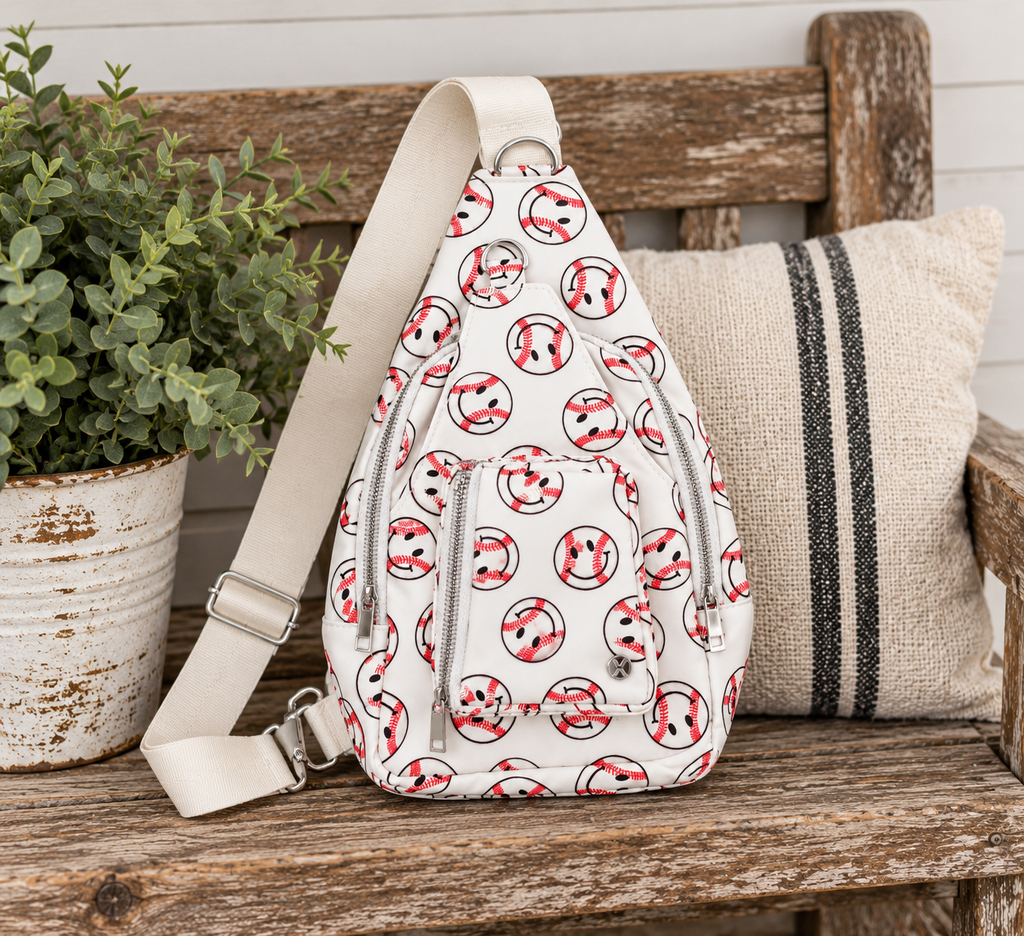 Baseball Smiley Sling Bag displayed on rustic wooden bench showing white all-over baseball smiley face print, two front silver zipper compartments, and wide flat cream adjustable strap beside distressed white clay pot with greenery and grain sack striped pillow in farmhouse setting
