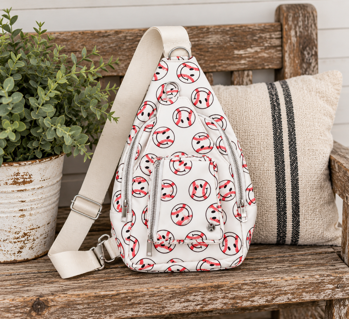 Baseball Smiley Sling Bag displayed on rustic wooden bench showing white all-over baseball smiley face print, two front silver zipper compartments, and wide flat cream adjustable strap beside distressed white clay pot with greenery and grain sack striped pillow in farmhouse setting
