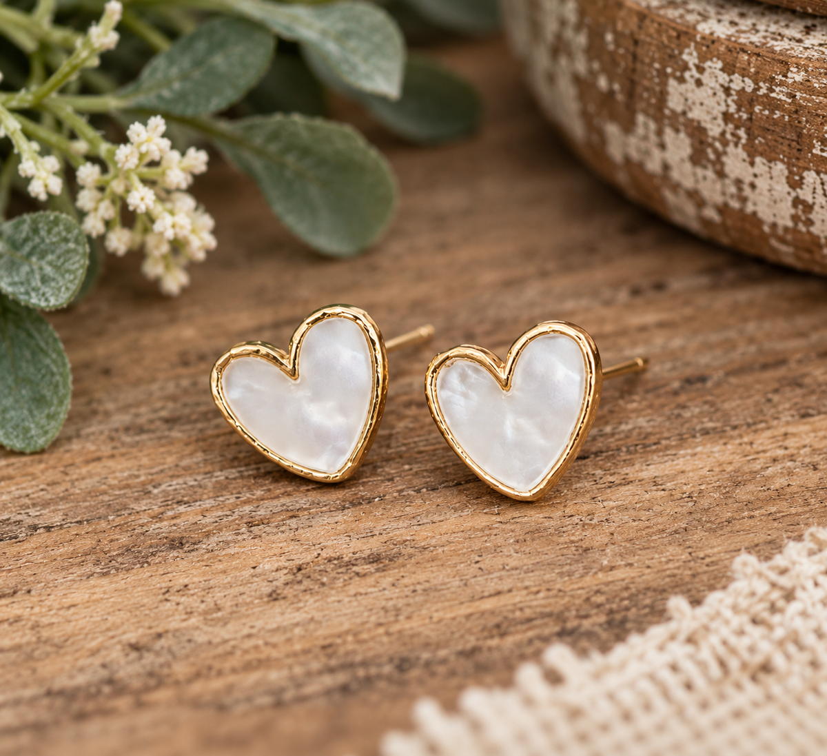 Pearl heart stud earrings with gold-tone trim displayed on rustic wood surface with baby's breath, eucalyptus, chippy white box, and cream lace textile
