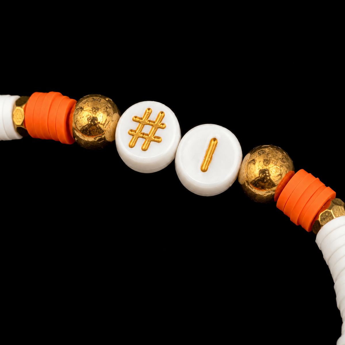 Close-up of white #1 letter beads with gold text flanked by textured gold-tone accent balls and orange and white disc beads on stretch bracelet

