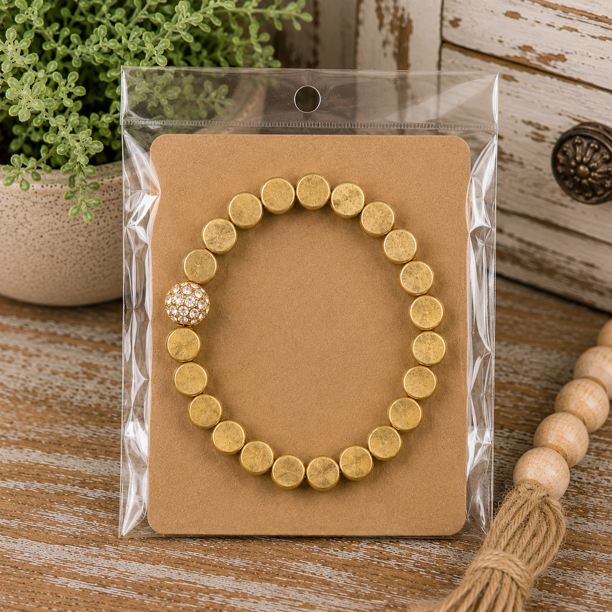 Gold crystal accent stretch bracelet on kraft card in clear packaging displayed on rustic wood surface with wood bead garland
