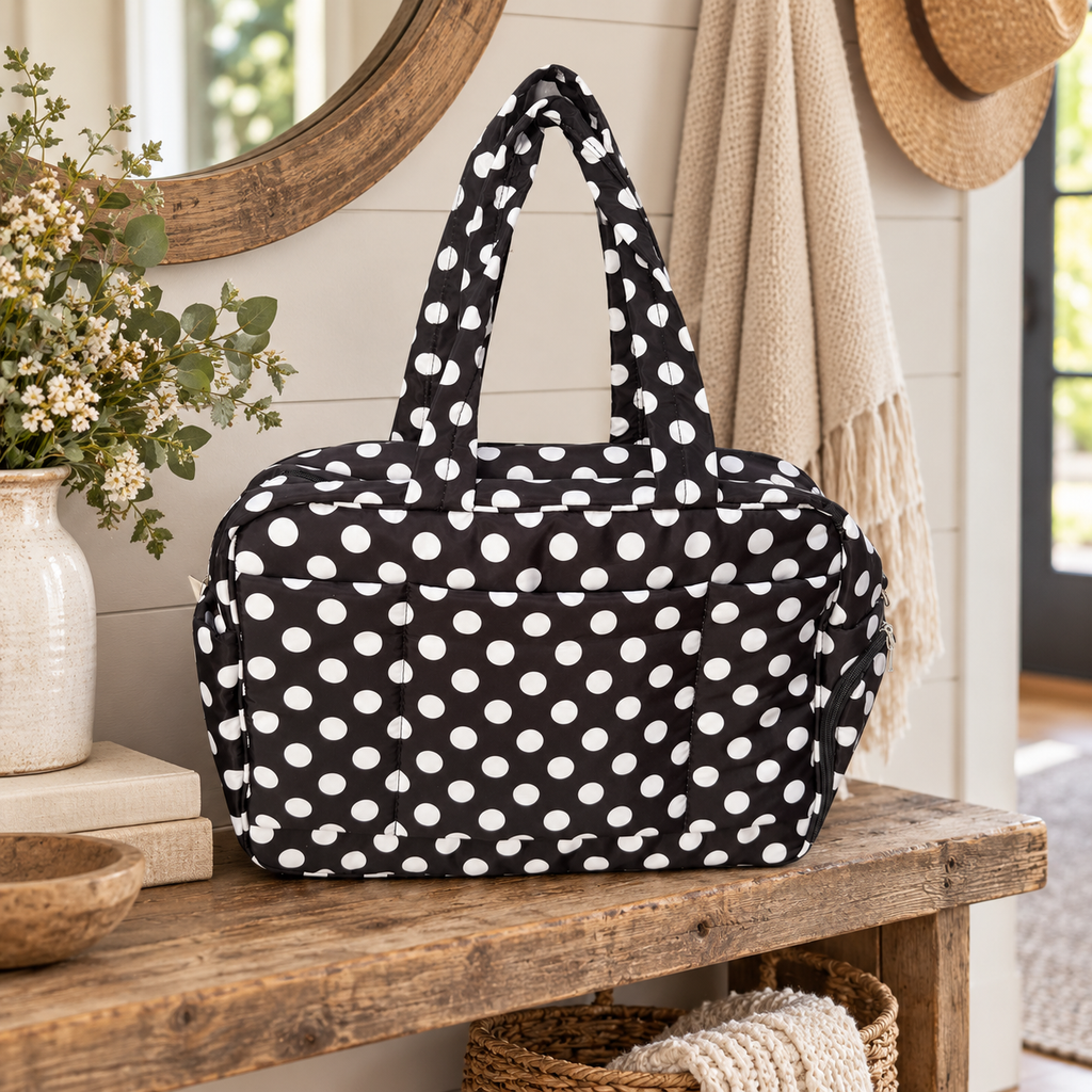 Black Polka Dot Shoulder Tote Bag on rustic wood entryway bench showing black padded fabric with bold white polka dots and dual shoulder handles beside cream stoneware vase of white wildflowers, stacked books, round wood mirror, and fringe throw with straw hat
