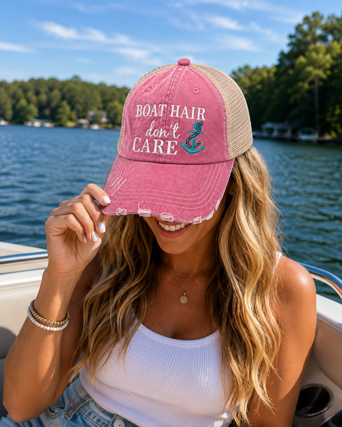 A woman on a boat wearing a distressed trucker hat with a pink front panel, beige mesh back, and embroidered text reading 'BOAT HAIR don't CARE' with a teal anchor graphic.