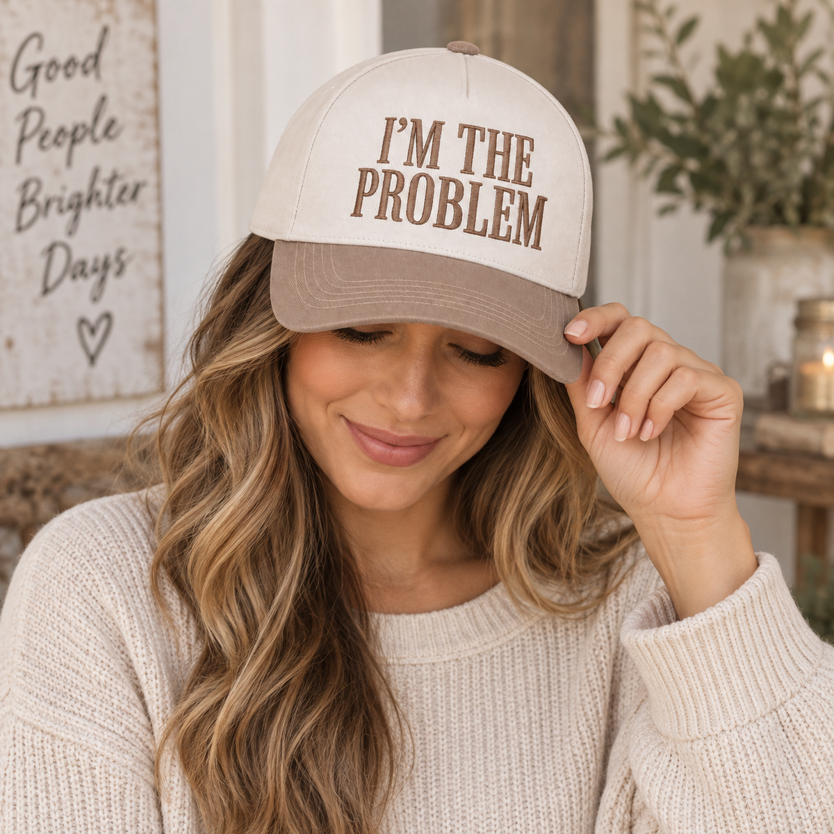 Brunette woman with long wavy highlighted hair in cream knit sweater smiling while touching brim of I'm the Problem Baseball Cap in Ash Mocha, in farmhouse interior with Good People Brighter Days sign and greenery in background
