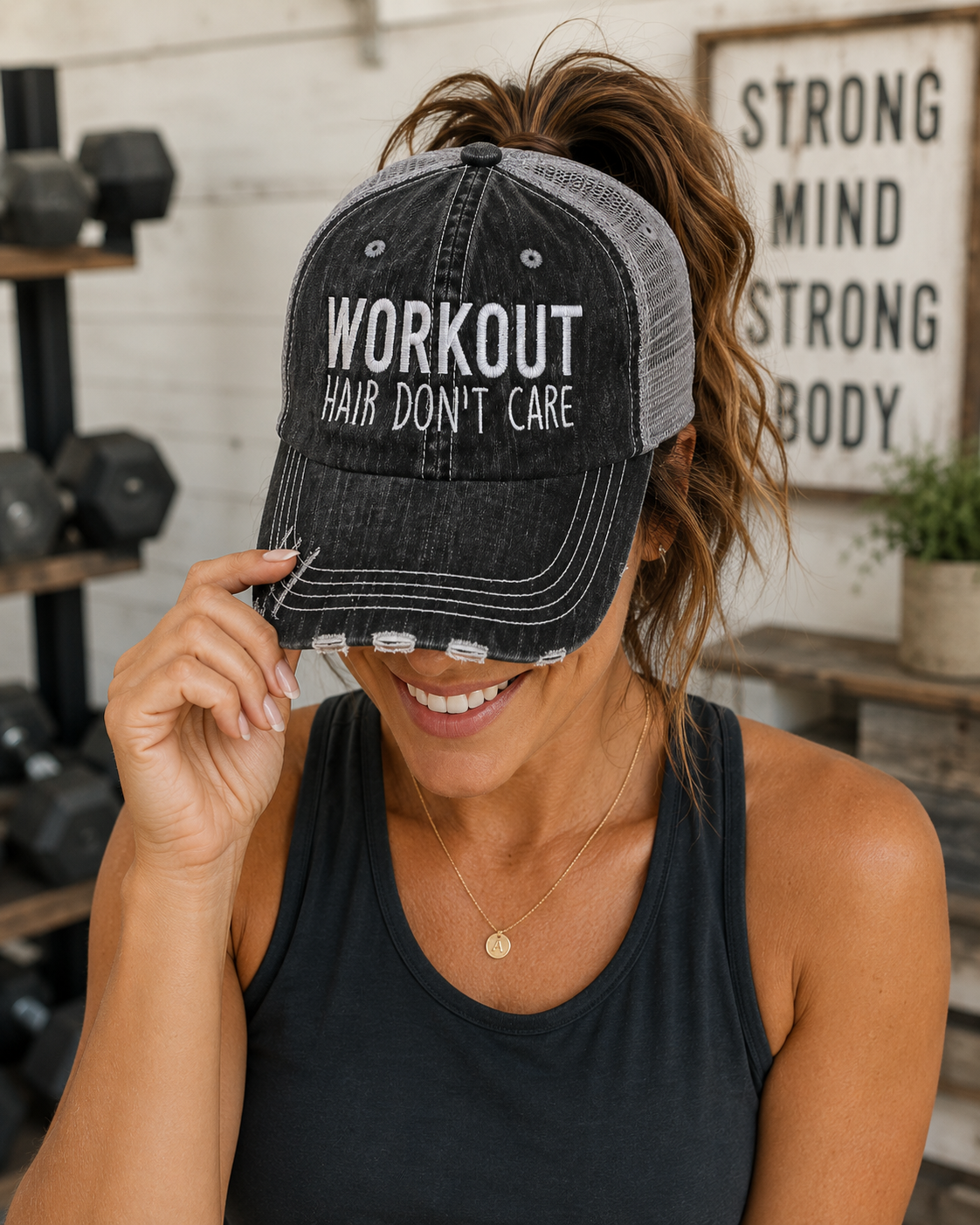 A woman wearing a distressed black trucker-style baseball cap with white text that reads 'WORKOUT HAIR DON'T CARE'. The cap has a washed black denim-look front panel and a gray mesh back.