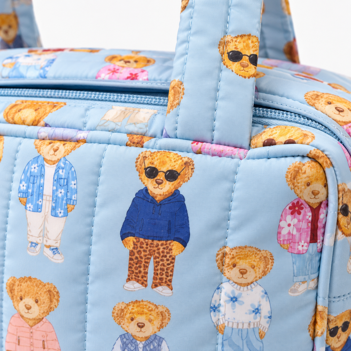 Close-up of Blue Teddy Bear Weekender Bag showing quilted channel stitching on light blue nylon with allover teddy bear print featuring bears in navy hoodie with leopard pants, pink floral jacket, white knit sweater, and blue floral shirt with sunglasses, and dual padded handle attachment
