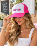 Woman wearing Good Vibes Trucker Hat with white front and hot pink bill and mesh back tilted down showing multicolor Good Vibes embroidery with smiley face detail and Good Days Ahead chalkboard backdrop
