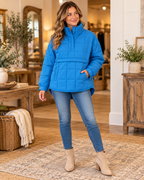 Woman wearing a bright blue quilted puffer pullover jacket with jeans and tan ankle boots in a boutique setting

