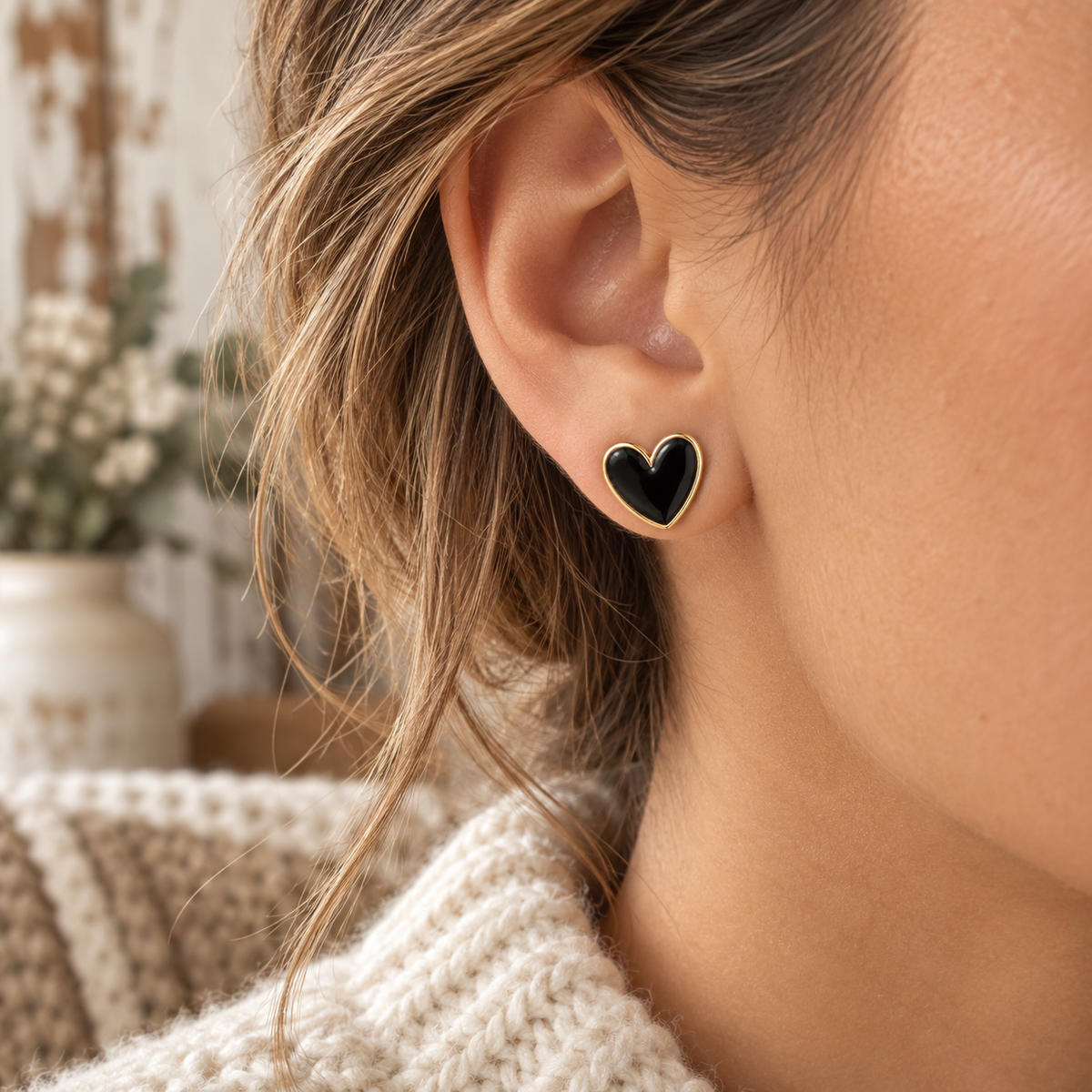 Woman wearing black heart stud earring with gold-tone trim on ear with highlighted hair, cream chunky knit sweater, and white floral backdrop
