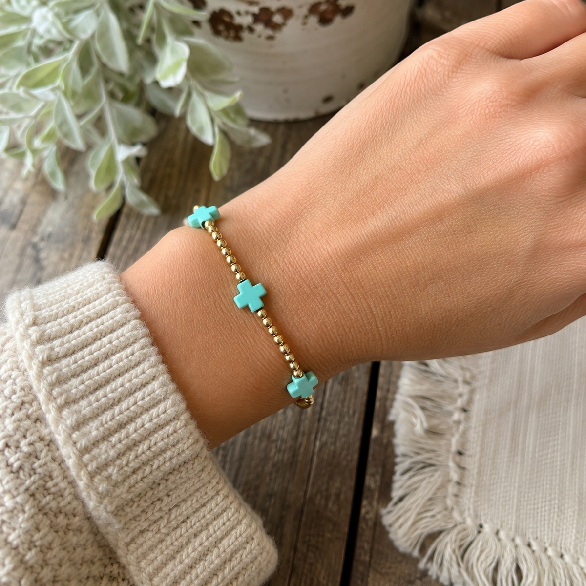 Woman wearing gold beaded stretch bracelet with turquoise cross accents on wrist with cream knit sweater, rustic wood surface, and chippy white pot with eucalyptus
