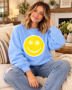 Seated model wearing a light blue sweatshirt with a yellow happy face and white daisy eyes, paired with jeans
