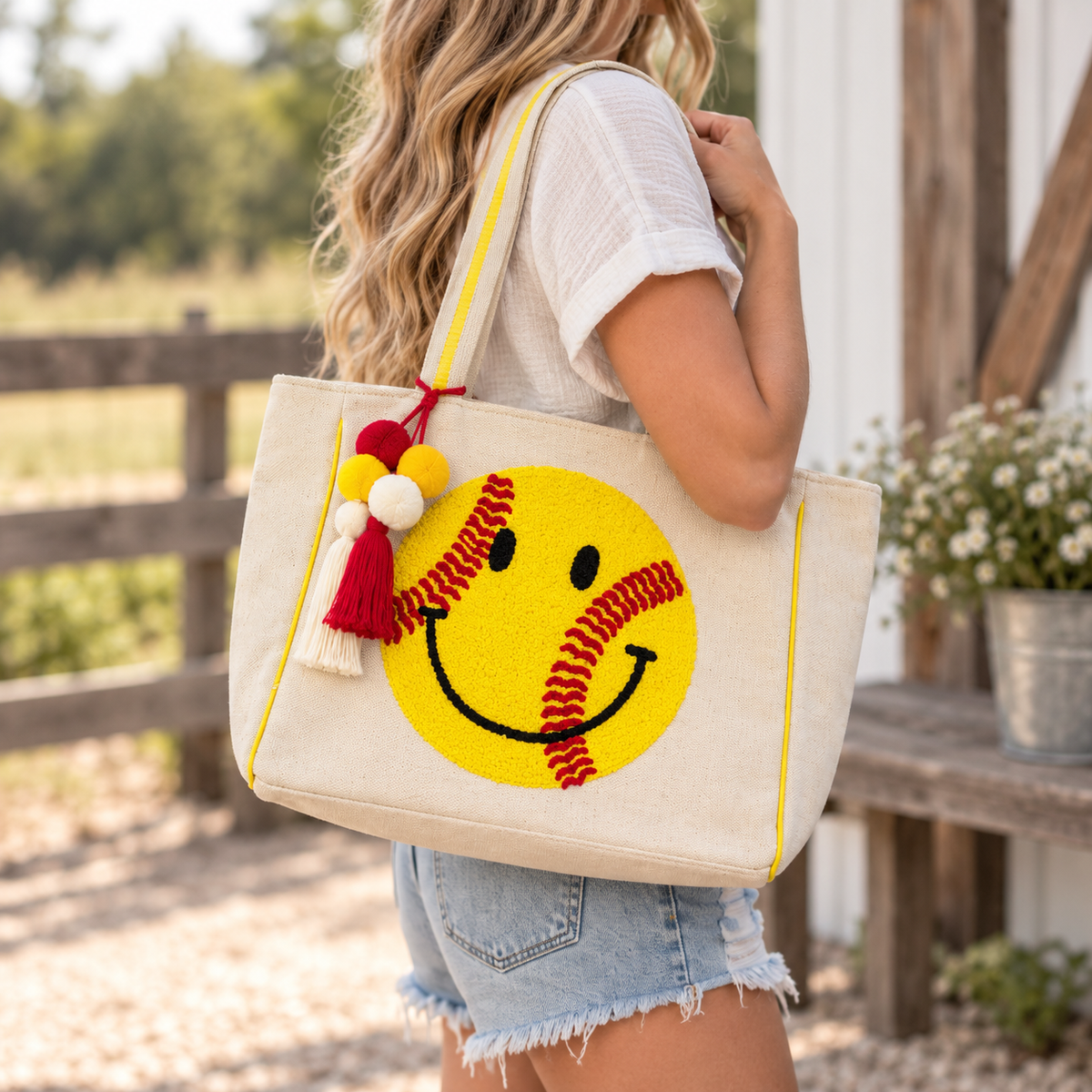 Woman carrying Softball Smiley Tote Bag over shoulder showing natural canvas body with large yellow textured softball smiley face, red stitching, yellow trim, and colorful pom-pom tassel with white tee and farm backdrop
