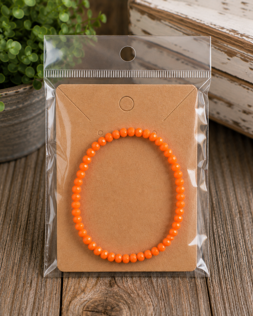 Orange faceted bead stretch bracelet on kraft card in clear packaging displayed on rustic wood surface with potted greenery and chippy white frame
