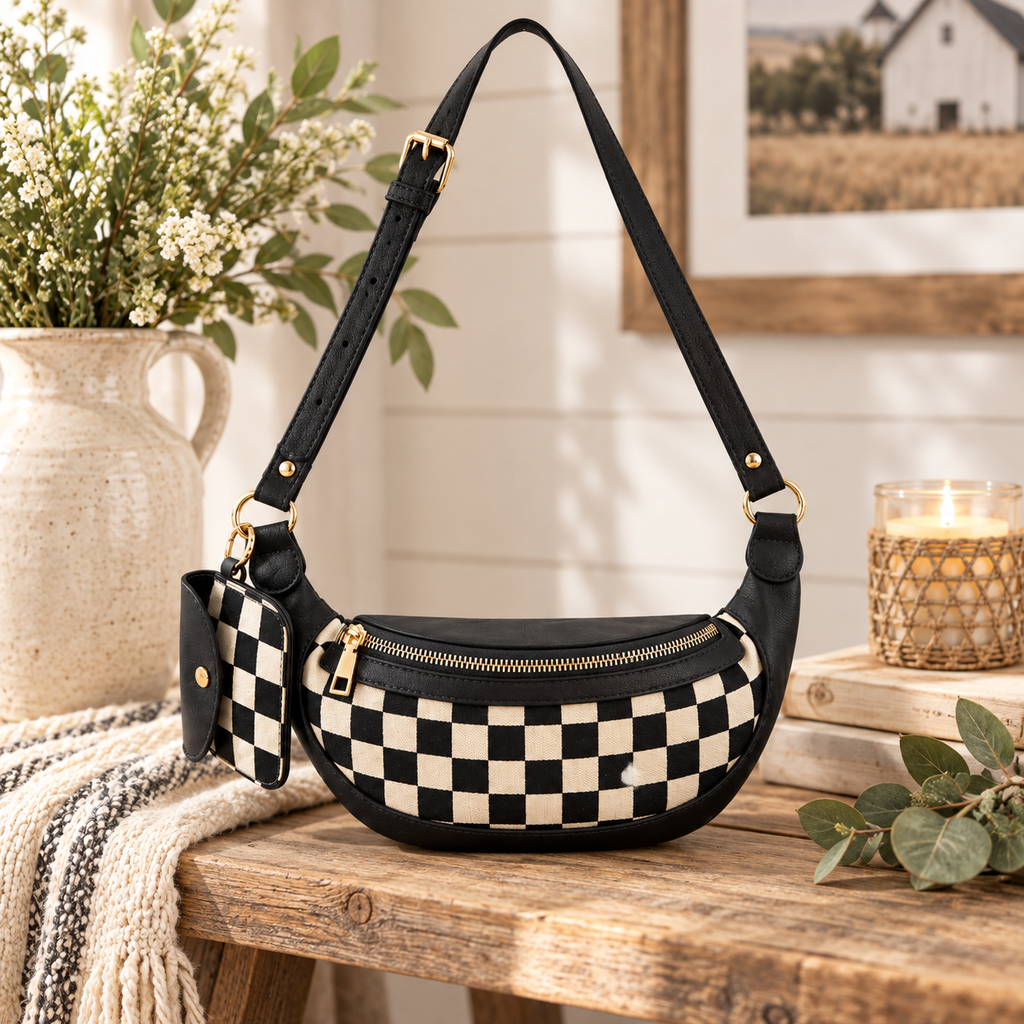 Duo Checkered Crossbody Bag in Black on rustic wood bench showing black and cream checkered body with black trim and gold zipper beside white ceramic jug with white florals, stacked books, grey stripe linen throw, and wicker candle holder on white shiplap background with barn painting on wall
