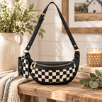 Duo Checkered Crossbody Bag in Black on rustic wood bench showing black and cream checkered body with black trim and gold zipper beside white ceramic jug with white florals, stacked books, grey stripe linen throw, and wicker candle holder on white shiplap background with barn painting on wall
