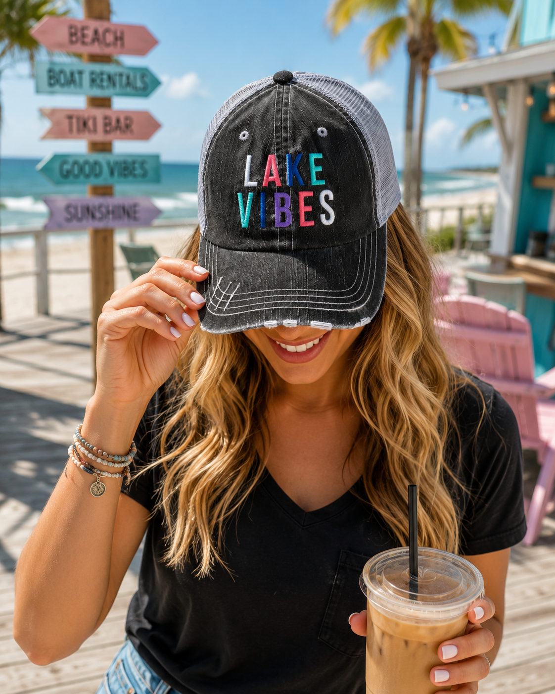 A woman wearing a black distressed trucker hat with 'LAKE VIBES' embroidered in colorful letters, standing on a beach boardwalk with a drink in hand.