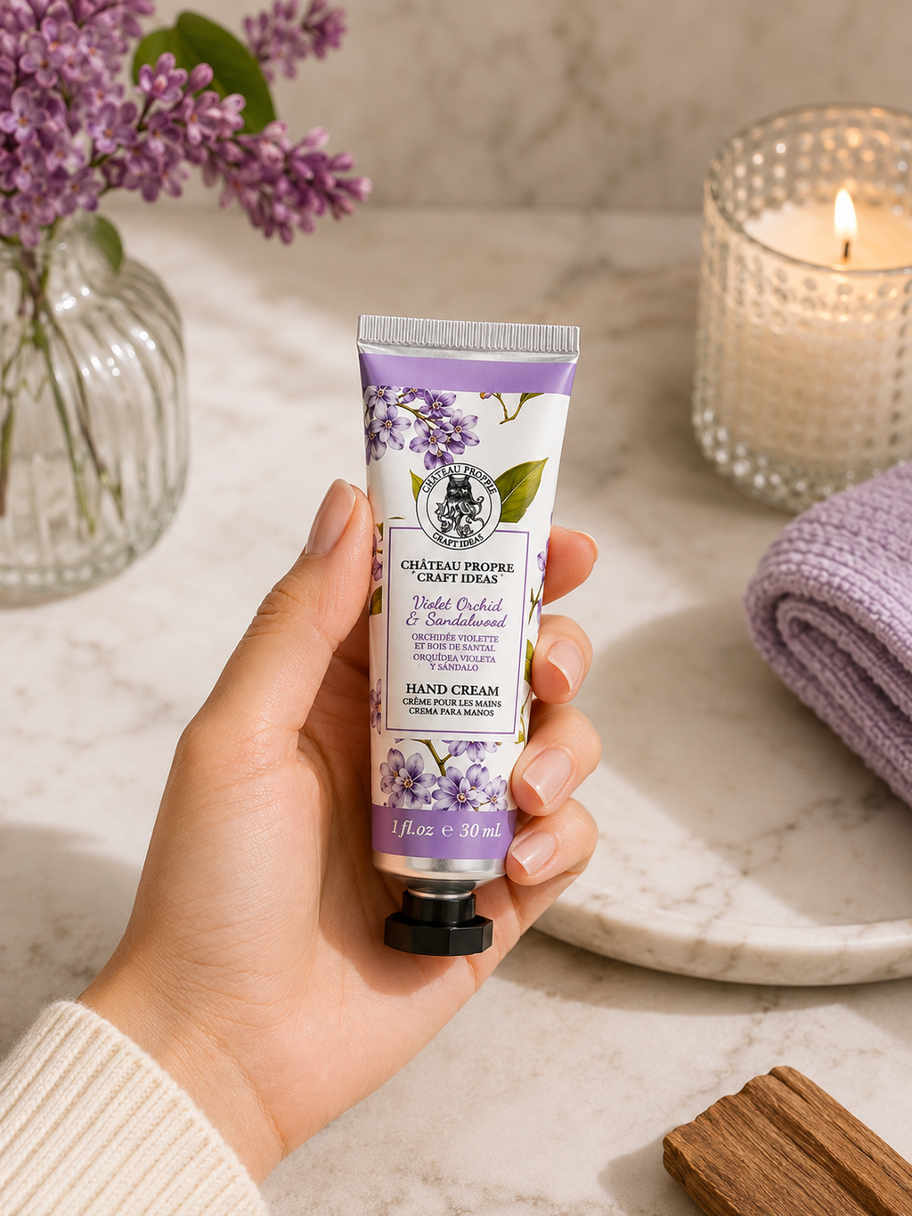 A hand holds a tube of Château Propre Craft Ideas hand cream with floral illustrations on the label, set against a spa-like background with a lit candle, flowers, and a soft towel.

