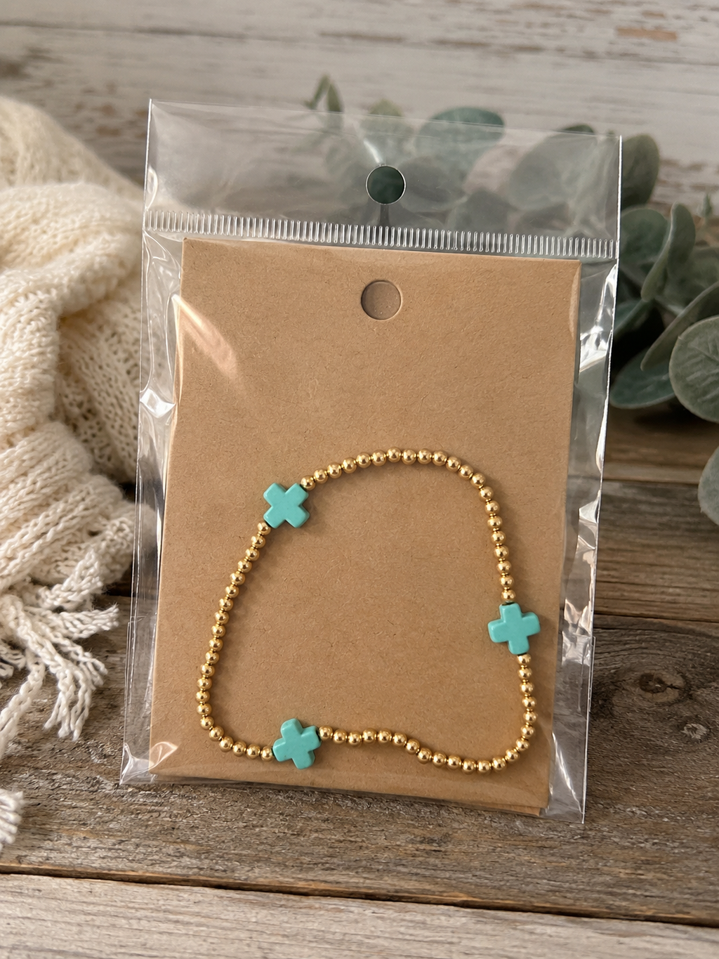 Gold beaded bracelet with turquoise cross accents on kraft card in clear packaging on rustic wood surface with cream knit scarf and eucalyptus greenery
