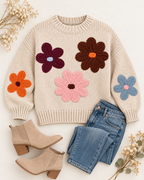 Tan floral knit sweater laid flat beside blue jeans and tan ankle boots, showing colorful flowers across the front and sleeves
