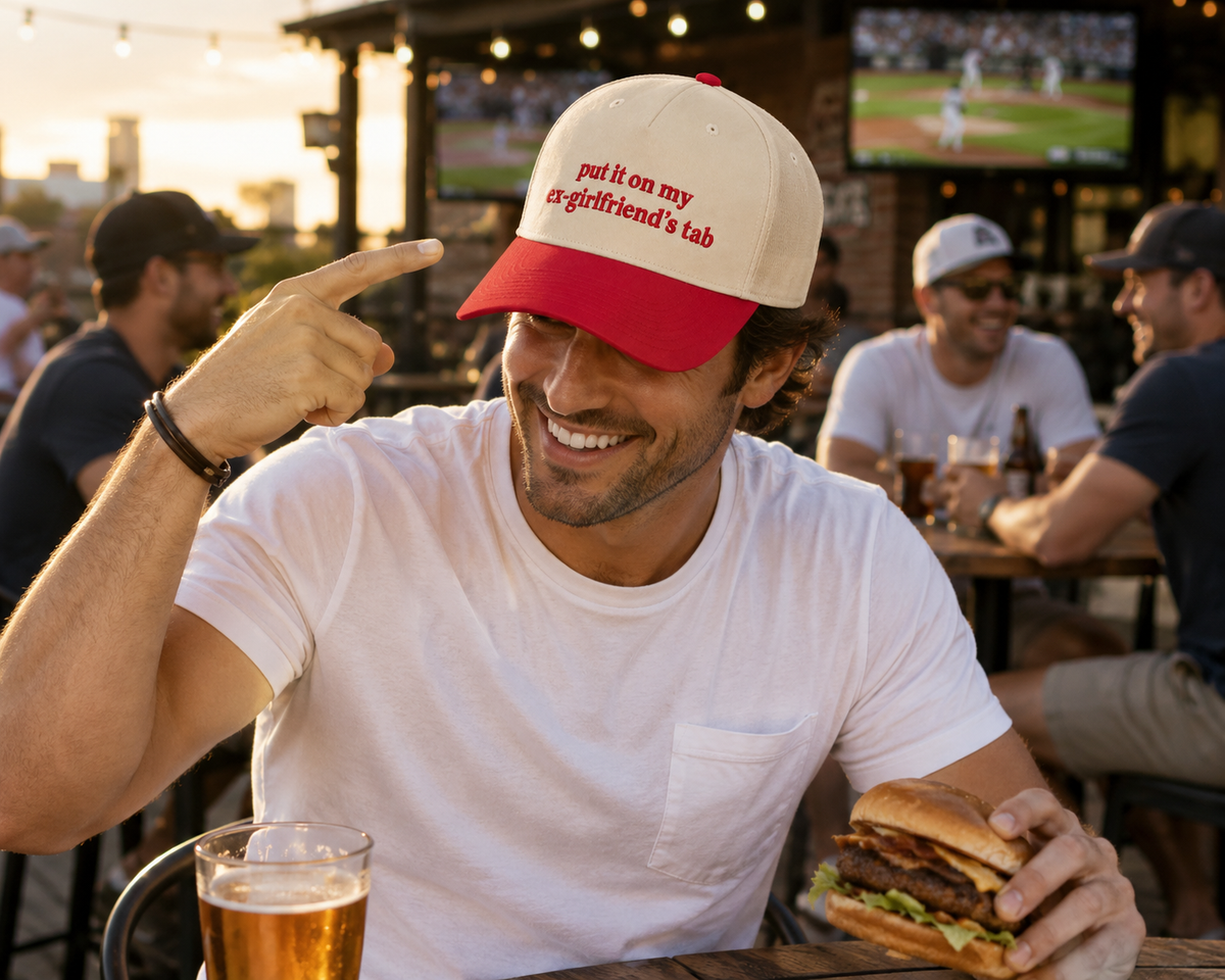 A man at an outdoor bar wearing a cream and red baseball cap that reads "Put It On My Ex-Girlfriend's Tab," holding a burger and pointing to the brim, with a beer in the foreground.
