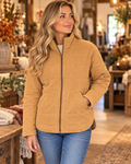 Blonde woman smiling in camel garment washed quilted jacket zipped up with blue jeans, in warm boutique interior with fall florals and white pumpkins
