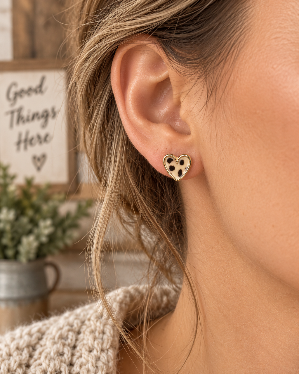 Woman wearing leopard heart stud earring with gold-tone trim on ear with blonde hair, cream knit sweater, and Good Things Here wood sign backdrop
