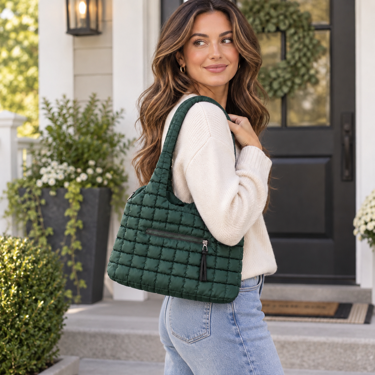Woman in cream sweater and jeans carrying Forest Green Quilted Tote Bag on shoulder showing forest green grid-quilted puffer nylon, single looped quilted shoulder strap, and exterior zippered front pocket on white farmhouse porch with dark greenery planters and black front door with wreath
