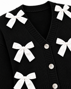 Close-up of Black Satin Bow Knit Cardigan showing deep V-neckline with ribbed trim, pearl dome buttons along button placket, and white satin ribbon bow detail on black ribbed knit on white background
