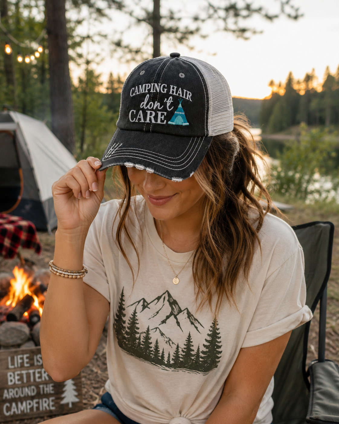 A woman at a campsite wearing a black and gray trucker hat with the text 'CAMPING HAIR don't CARE' and a small turquoise teepee graphic, paired with a beige graphic t-shirt.