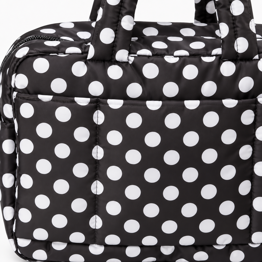 Close-up of Black Polka Dot Shoulder Tote Bag showing black padded quilted fabric texture with bold white polka dots, top zipper closure, and large front slip pocket detail on white background
