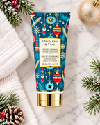 Frosted Coconut hand and body lotion laid flat beside white towels, frosted evergreen branches, pinecones, and a gold Christmas ornament
