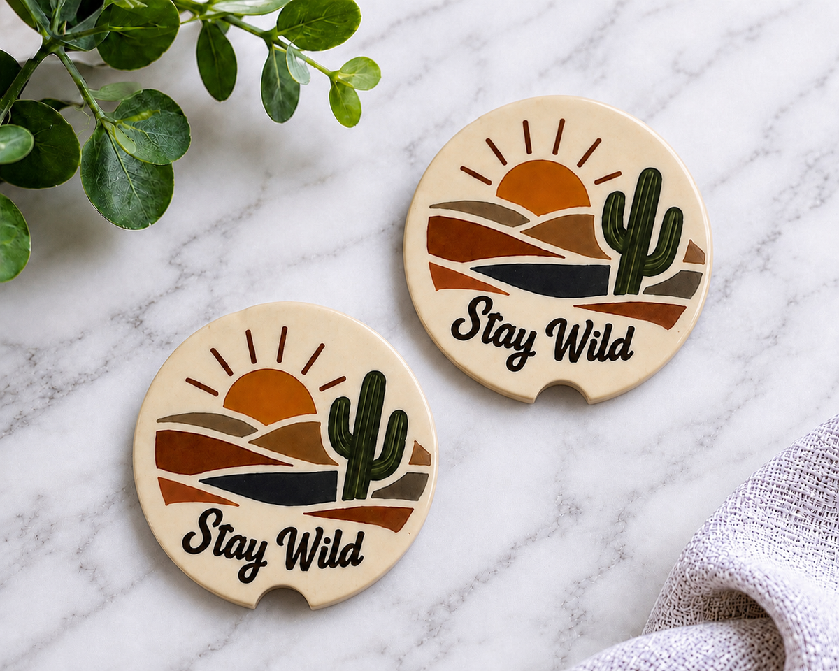 Stay Wild Car Coaster Set of 2 on a marble surface with greenery, showing desert sunset and cactus design on cream ceramic coasters with finger notch.
