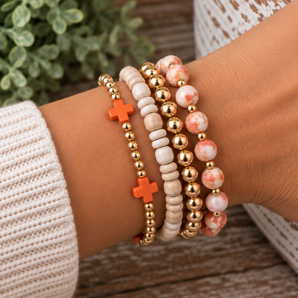 Gold and orange cross bracelet stacked with white heishi gold bead bracelet and peach marble stone gold bead bracelet on wrist with white knit sleeve, rustic wood surface, and greenery backdrop
