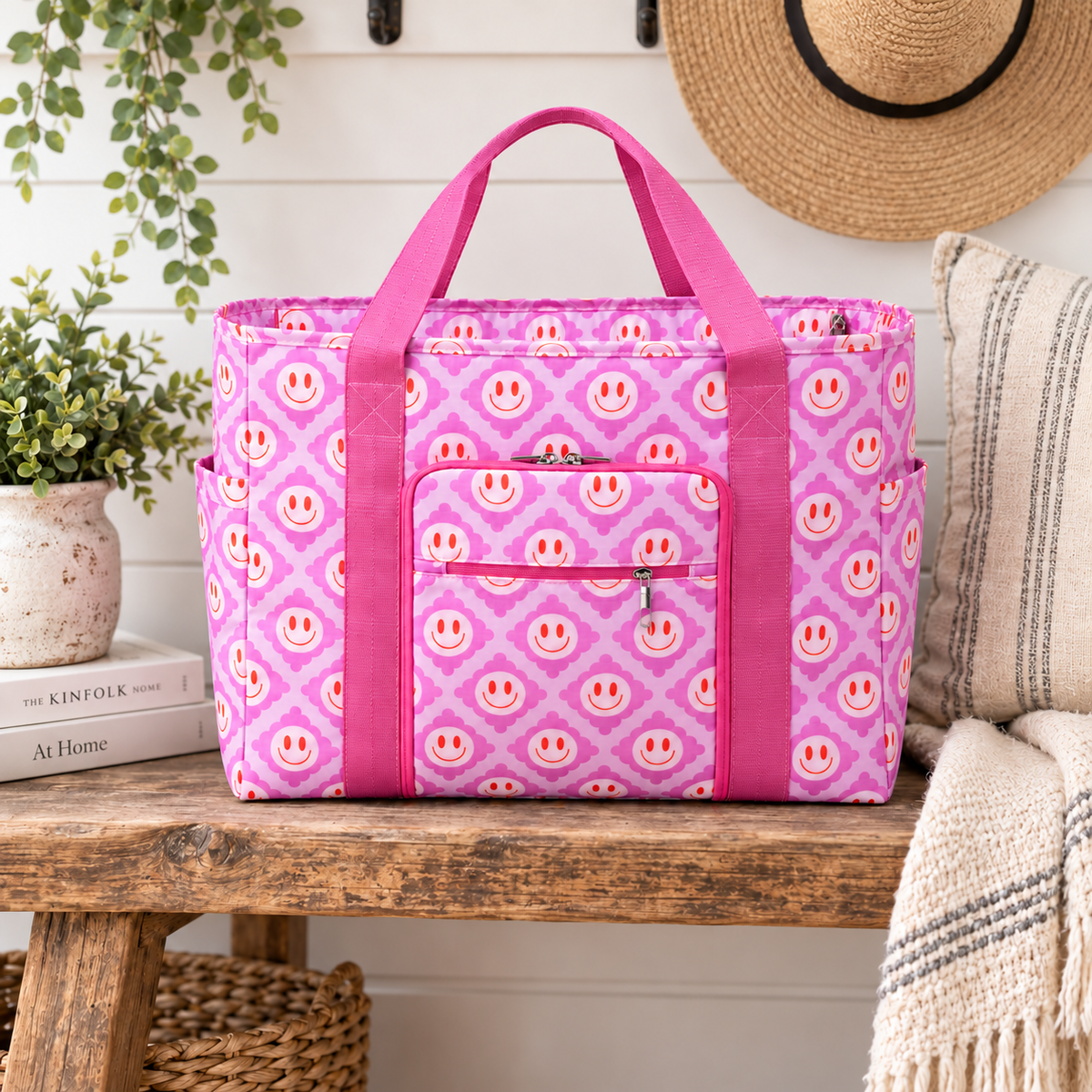 Pink Smiley Flower Weekender Bag on rustic wood bench showing light pink all-over smiley face quatrefoil print, hot pink webbed handles, front zippered pocket, and side slip pockets against white shiplap wall with cream potted greenery, The Kinfolk Home and At Home books, straw hat, and striped throw
