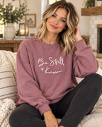 Model wearing the dusty mauve Be Still + Know crewneck sweatshirt with white script lettering and black pants
