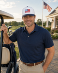 A smiling man in a navy blue polo shirt and khaki pants stands next to a golf cart on a golf course, wearing a white and navy baseball cap with an American flag graphic.