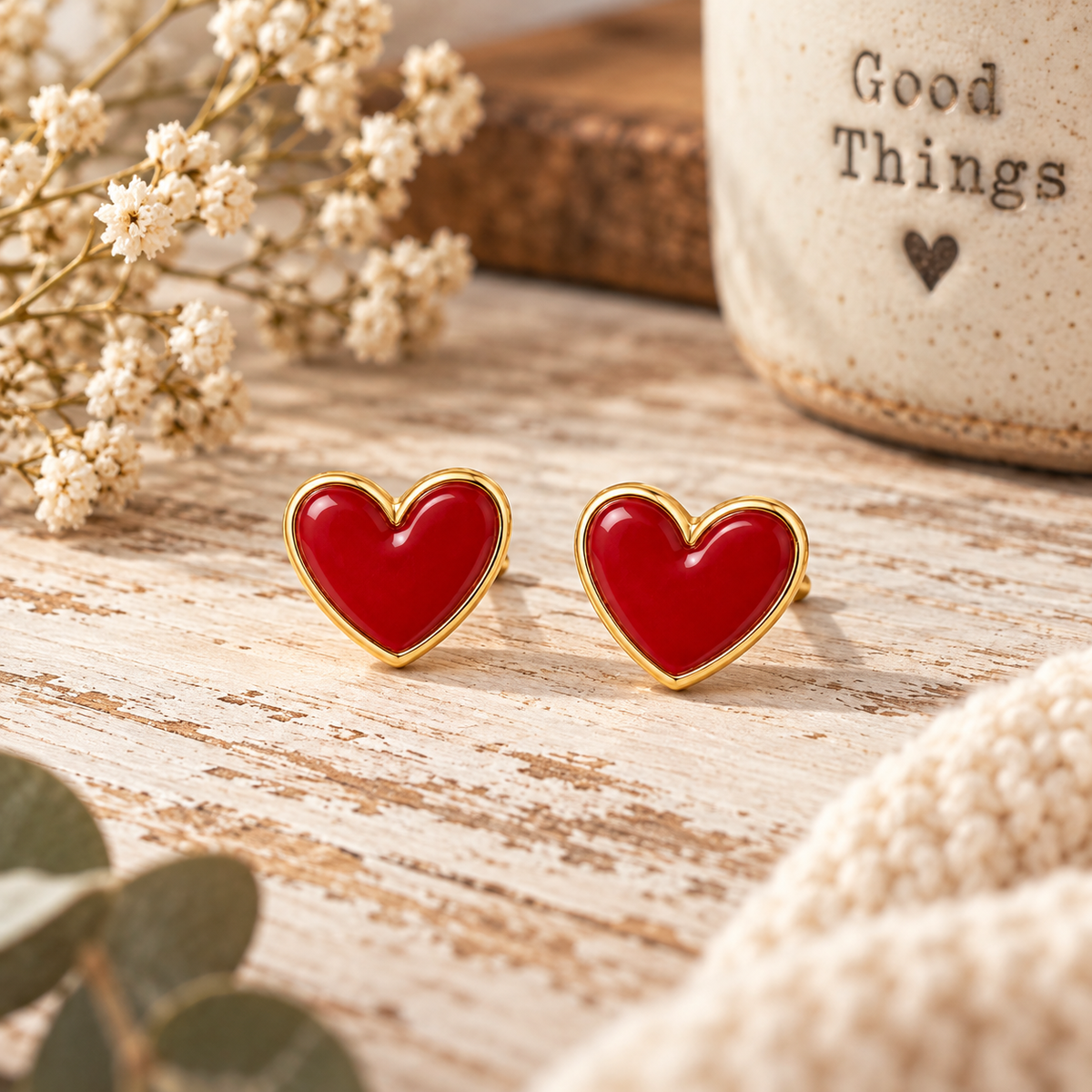 Red heart stud earrings with gold-tone trim displayed on chippy white wood surface with Good Things stoneware mug, baby's breath, eucalyptus, and cream knit
